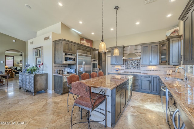 kitchen with arched walkways, stainless steel appliances, a sink, light stone countertops, and wall chimney exhaust hood