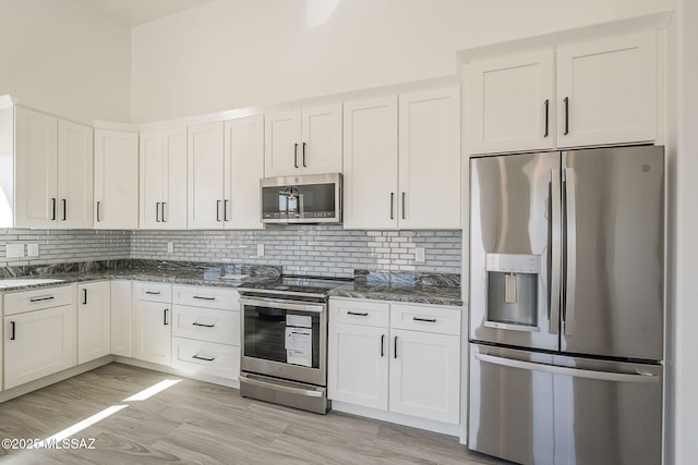 kitchen featuring light wood finished floors, tasteful backsplash, appliances with stainless steel finishes, white cabinetry, and dark stone countertops