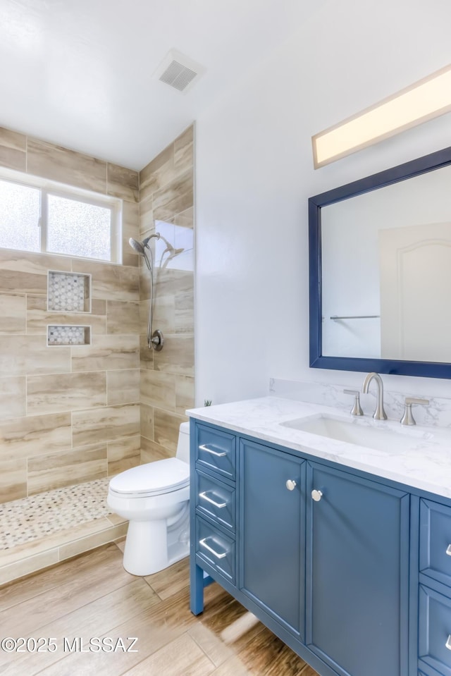 bathroom with visible vents, toilet, wood finished floors, tiled shower, and vanity