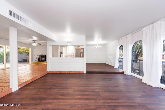 unfurnished living room featuring ceiling fan with notable chandelier, brick wall, dark wood finished floors, and visible vents