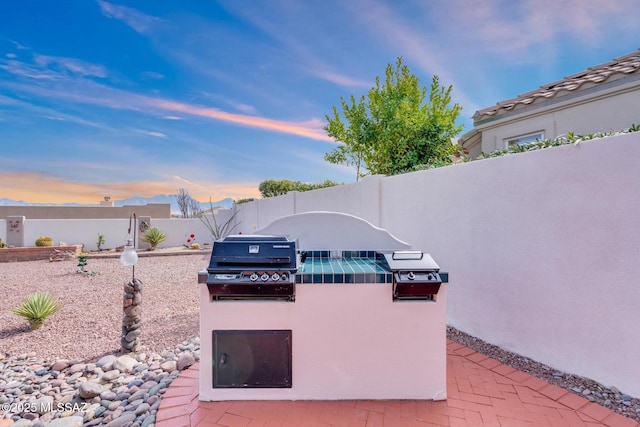 patio terrace at dusk featuring an outdoor kitchen, a fenced backyard, and a grill