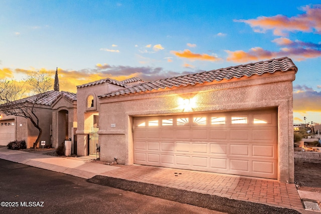 mediterranean / spanish-style home featuring stucco siding, a tile roof, a gate, decorative driveway, and a garage