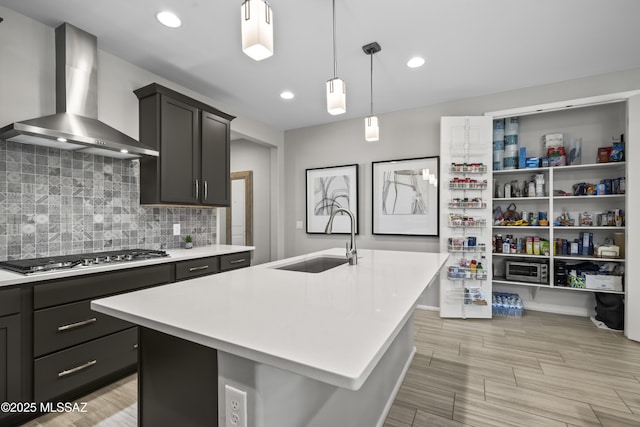 kitchen featuring a kitchen island with sink, a sink, light countertops, wall chimney exhaust hood, and stainless steel gas stovetop