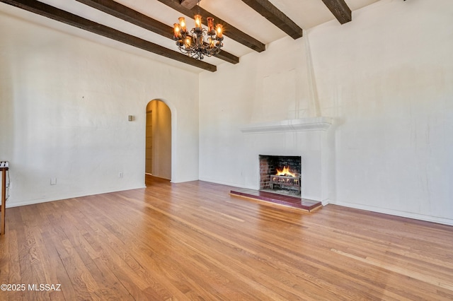 unfurnished living room with arched walkways, a warm lit fireplace, wood finished floors, beam ceiling, and an inviting chandelier