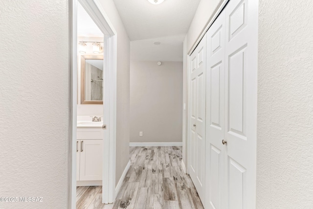 hallway featuring light wood finished floors, a sink, baseboards, and a textured wall