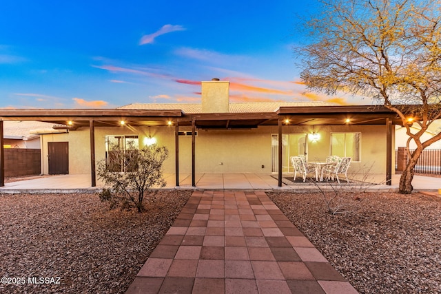 back of property at dusk with a patio area, stucco siding, a chimney, and fence