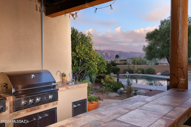 patio terrace at dusk with a fenced in pool, an outdoor kitchen, area for grilling, a mountain view, and fence