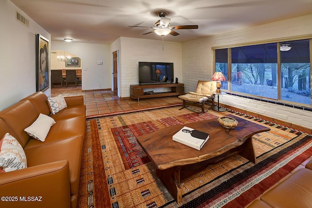 tiled living room featuring ceiling fan with notable chandelier, visible vents, and brick wall
