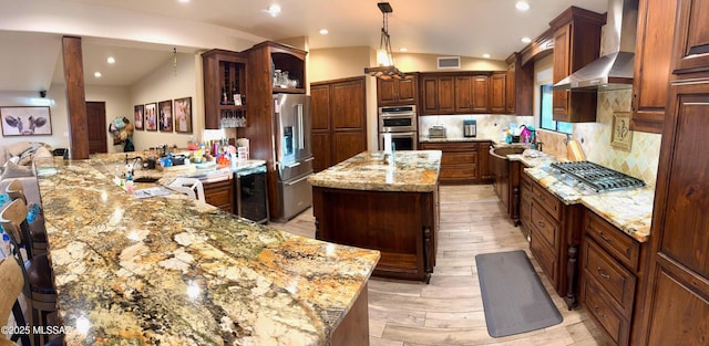 kitchen featuring lofted ceiling, light stone counters, stainless steel appliances, wall chimney range hood, and a center island