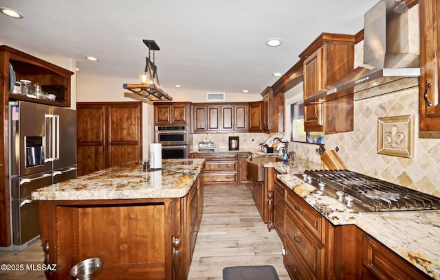 kitchen featuring light stone counters, a sink, a kitchen island, appliances with stainless steel finishes, and wall chimney range hood