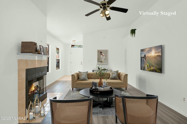 living area with a tiled fireplace, ceiling fan, vaulted ceiling, wood finished floors, and baseboards