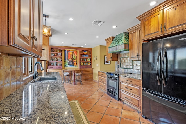 kitchen with light tile patterned floors, dark stone counters, custom range hood, black appliances, and a sink