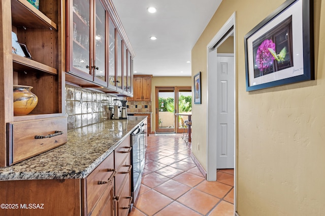 kitchen with light tile patterned floors, tasteful backsplash, glass insert cabinets, brown cabinets, and light stone counters