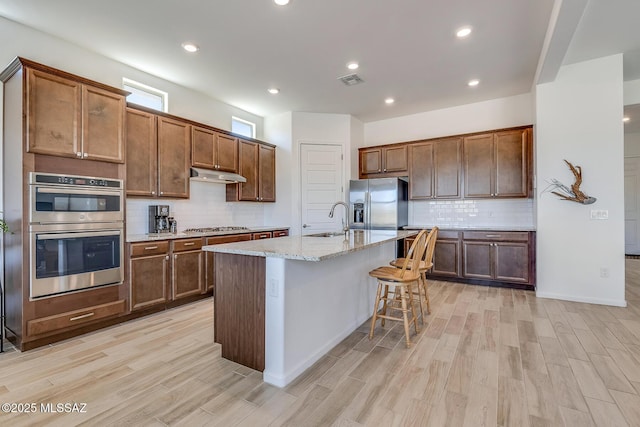 kitchen with light wood finished floors, an island with sink, a sink, under cabinet range hood, and appliances with stainless steel finishes