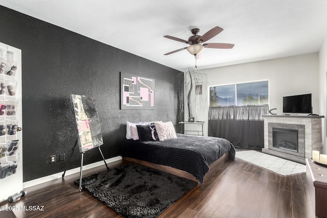 bedroom with baseboards, wood finished floors, a tile fireplace, a textured wall, and a ceiling fan