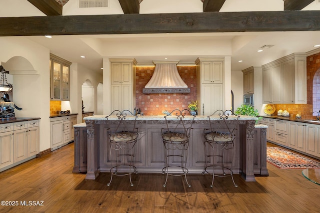 kitchen featuring wood finished floors, custom exhaust hood, beam ceiling, decorative backsplash, and a large island with sink