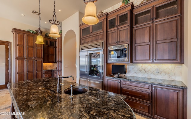 kitchen featuring an island with sink, a sink, arched walkways, built in appliances, and hanging light fixtures