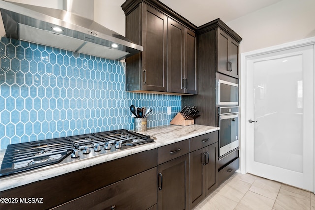 kitchen featuring decorative backsplash, wall chimney exhaust hood, light stone counters, stainless steel appliances, and dark brown cabinets