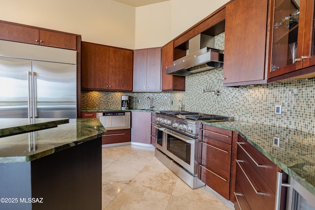 kitchen with dark stone countertops, tasteful backsplash, wall chimney exhaust hood, glass insert cabinets, and high end appliances