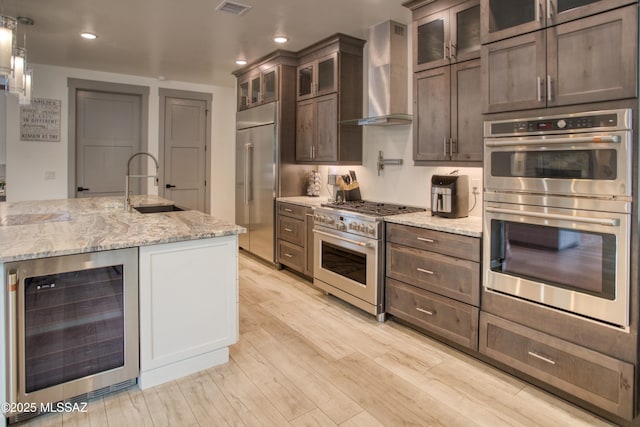 kitchen with dark brown cabinetry, a sink, high quality appliances, beverage cooler, and wall chimney exhaust hood