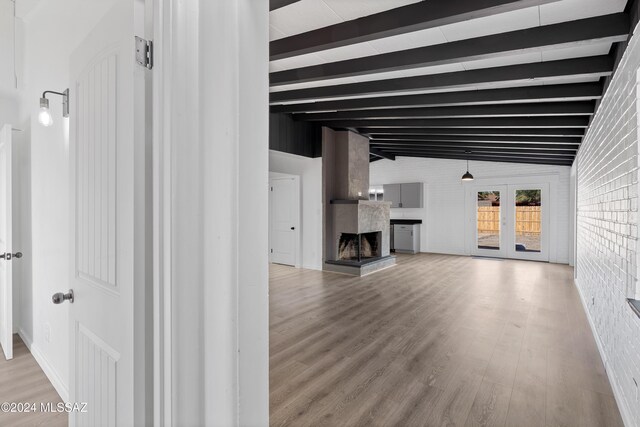 unfurnished living room featuring a fireplace with raised hearth, lofted ceiling with beams, brick wall, wood finished floors, and french doors