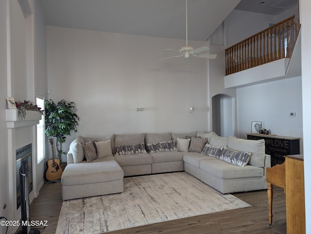 living area featuring wood finished floors, a ceiling fan, arched walkways, a towering ceiling, and a glass covered fireplace