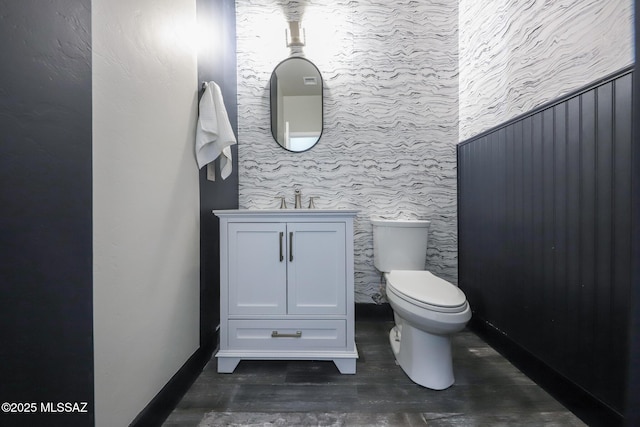 half bath featuring toilet, wood finished floors, wallpapered walls, baseboards, and vanity