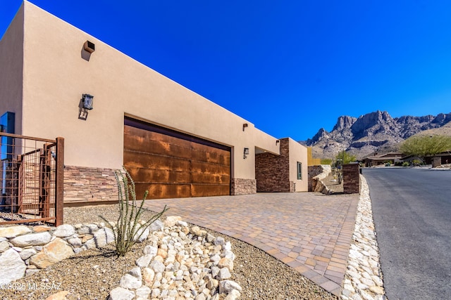 view of home's exterior featuring stucco siding, a garage, stone siding, decorative driveway, and a mountain view