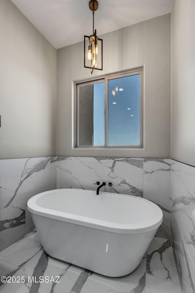 full bath featuring marble finish floor, a soaking tub, a notable chandelier, and tile walls