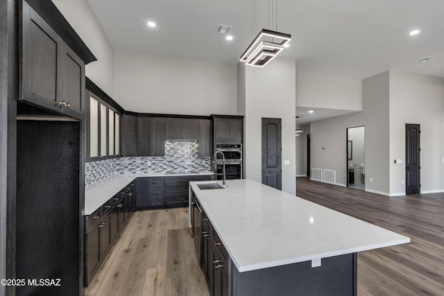 kitchen with an island with sink, stainless steel double oven, a high ceiling, light wood finished floors, and decorative backsplash