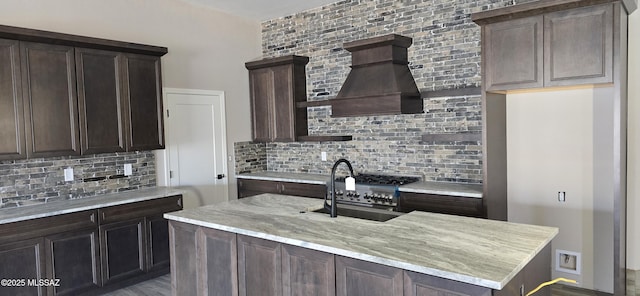 kitchen featuring dark brown cabinetry, premium range hood, a sink, decorative backsplash, and light stone countertops