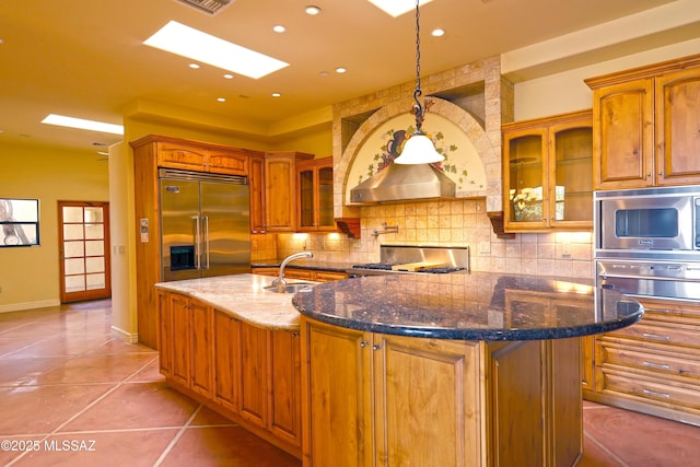 kitchen featuring light tile patterned floors, brown cabinetry, decorative backsplash, an island with sink, and built in appliances