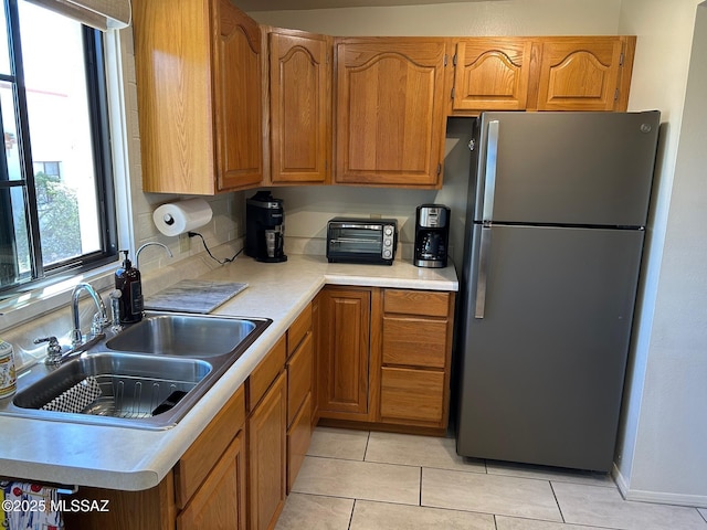 kitchen featuring light tile patterned floors, a sink, light countertops, freestanding refrigerator, and brown cabinetry