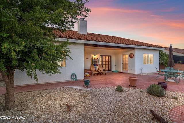 back of property at dusk with a tiled roof, a patio, a chimney, and stucco siding