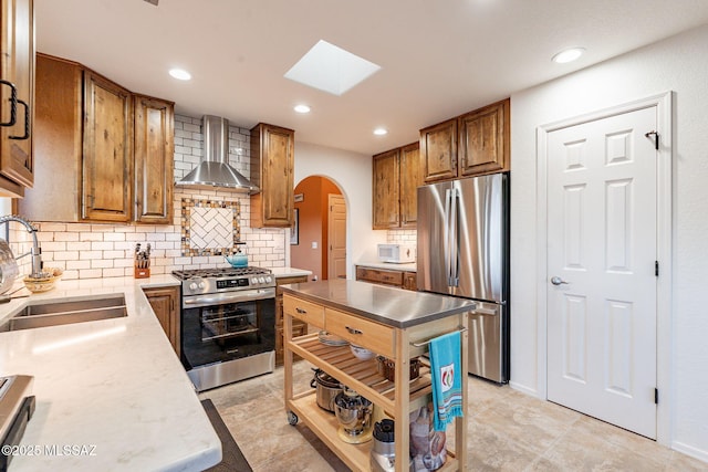 kitchen featuring arched walkways, stainless steel appliances, a sink, brown cabinets, and wall chimney exhaust hood