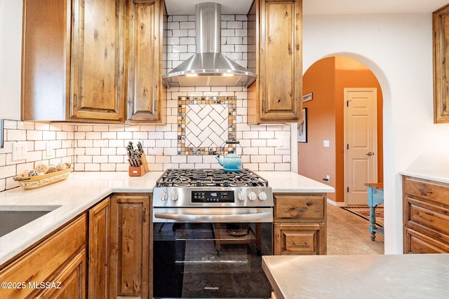 kitchen featuring arched walkways, decorative backsplash, brown cabinetry, stainless steel gas range, and wall chimney exhaust hood