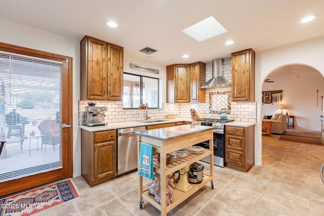 kitchen featuring visible vents, arched walkways, brown cabinetry, stainless steel appliances, and wall chimney range hood