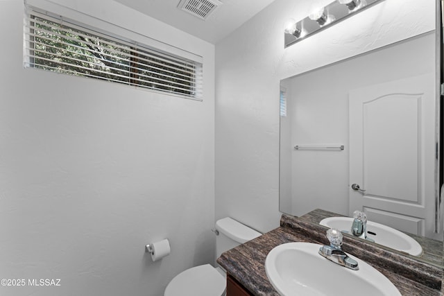 half bath with vanity, toilet, a textured wall, and visible vents