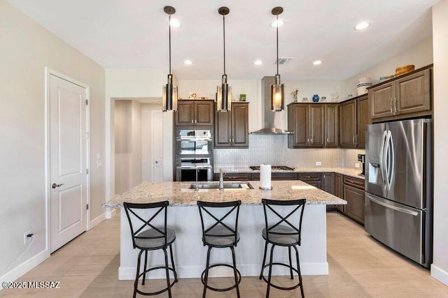 kitchen with a sink, backsplash, wall chimney range hood, stainless steel appliances, and a kitchen island with sink