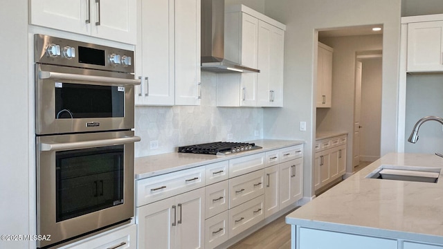 kitchen featuring a sink, tasteful backsplash, stainless steel appliances, wall chimney range hood, and light stone countertops