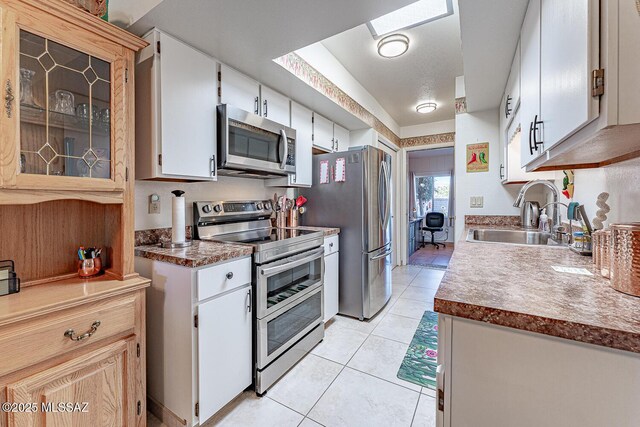 kitchen with appliances with stainless steel finishes, a skylight, light tile patterned flooring, white cabinets, and a sink