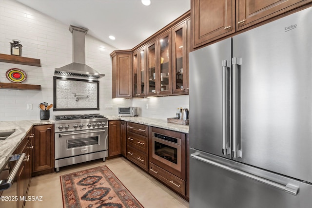 kitchen with premium appliances, backsplash, wall chimney exhaust hood, glass insert cabinets, and light stone countertops