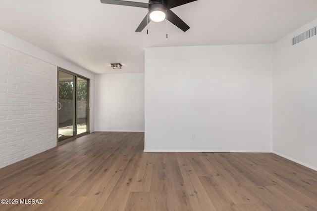 empty room with a ceiling fan, wood finished floors, visible vents, brick wall, and baseboards