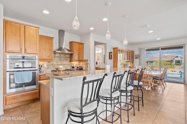 kitchen featuring light stone counters, tasteful backsplash, double oven, wall chimney range hood, and light tile patterned floors