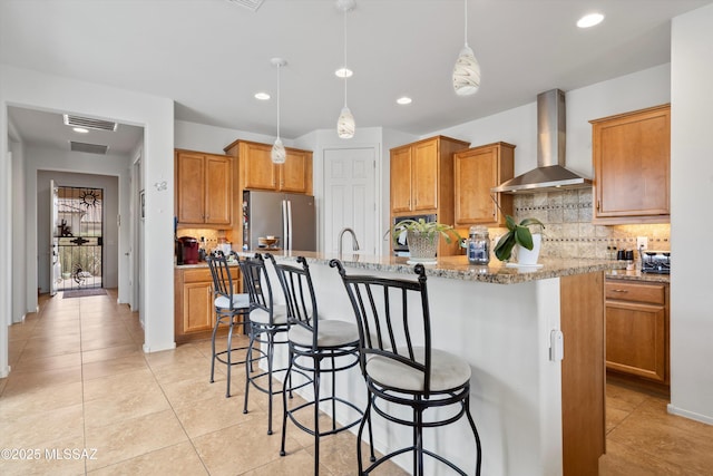 kitchen with tasteful backsplash, visible vents, freestanding refrigerator, wall chimney exhaust hood, and a kitchen island with sink