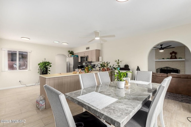 dining room featuring light wood-type flooring, a ceiling fan, arched walkways, baseboards, and a brick fireplace