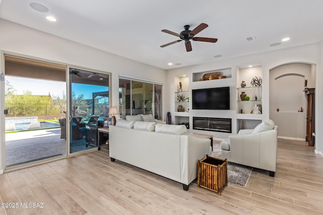 living room featuring visible vents, built in shelves, arched walkways, light wood finished floors, and ceiling fan