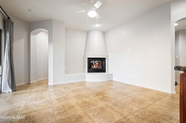 unfurnished living room featuring a ceiling fan, a large fireplace, arched walkways, light tile patterned flooring, and baseboards