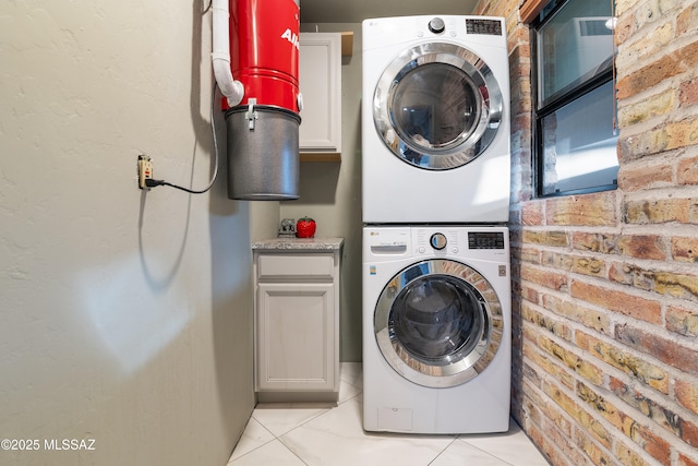 laundry area featuring light tile patterned floors, cabinet space, brick wall, and stacked washing maching and dryer