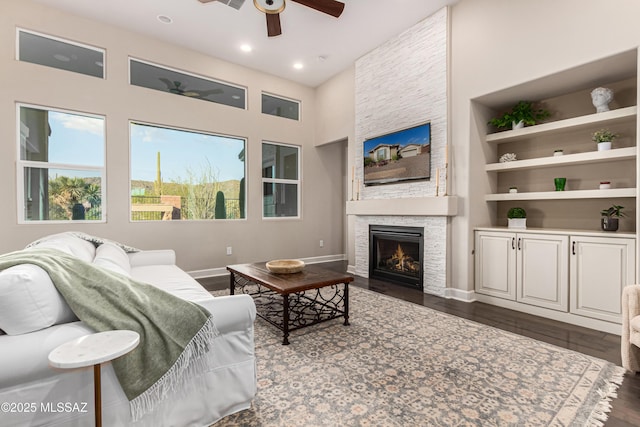 living room featuring built in features, dark wood finished floors, a fireplace, baseboards, and ceiling fan